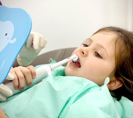 Niña sujetando un espejo mientras le cepillan los dientes con un cepillo eléctrico en la consulta de odontopediatría en Oviedo