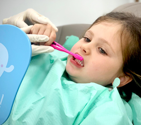 Niña aprendiendo a cepillarse con un cepillo manual mientras se mira en el espejo en la consulta infantil